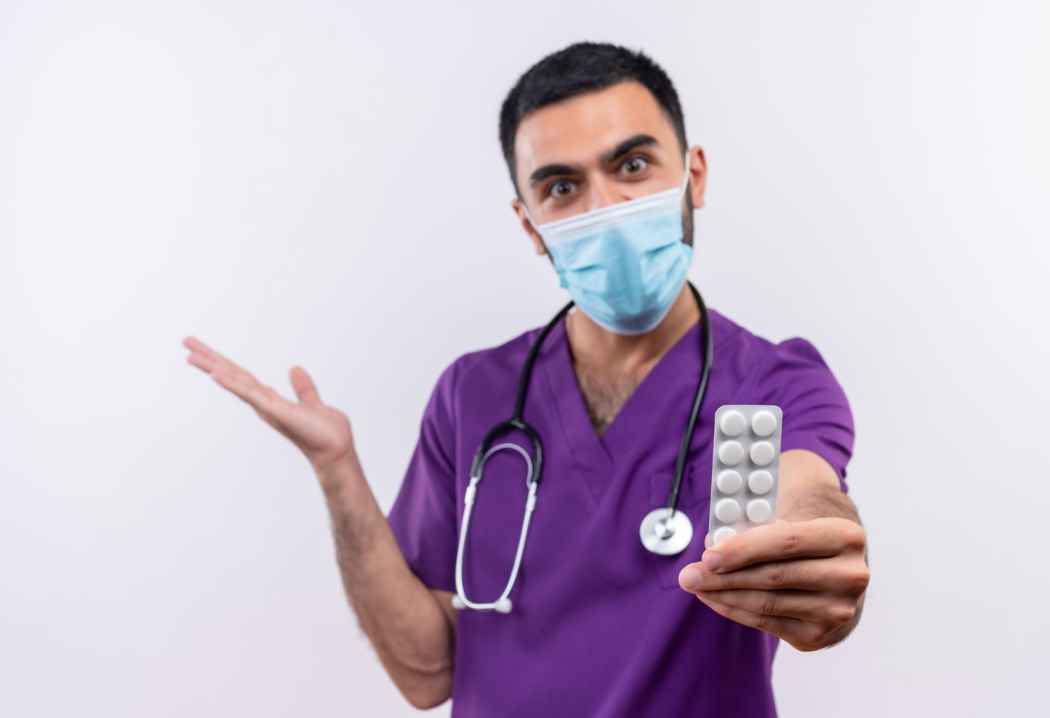 Doctor wearing scrubs and face mask holding a blister pack of tablets and explaining medication