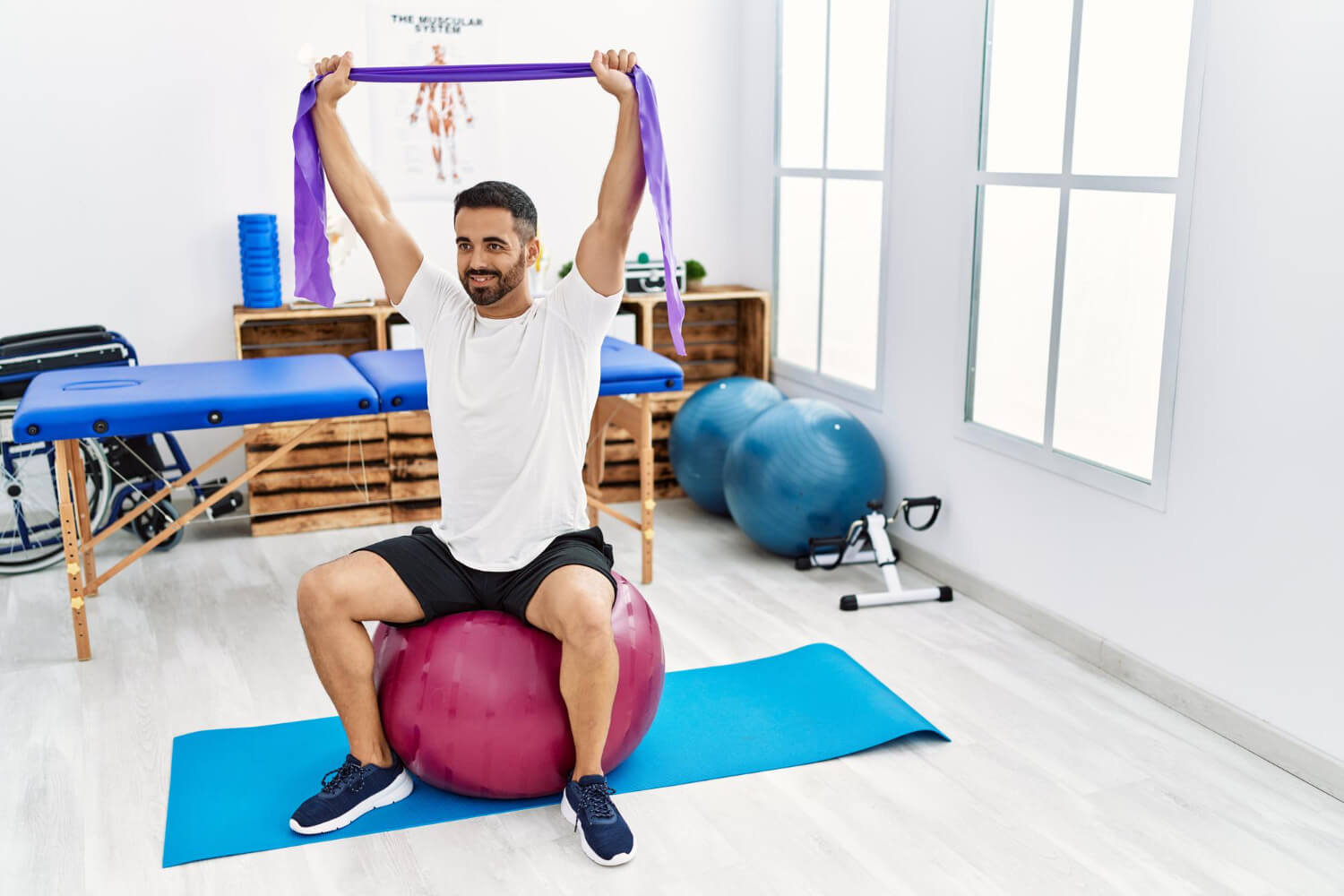 Man performing physiotherapy shoulder exercise using a resistance band while seated on a stability ball