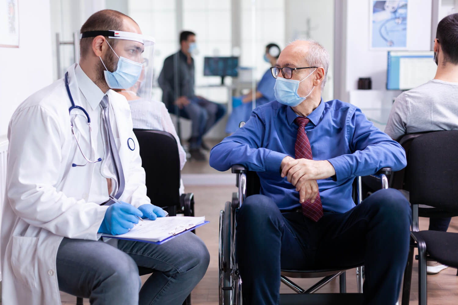Doctor wearing protective mask and face shield speaking with an elderly patient in a wheelchair