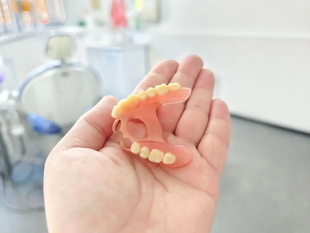 Hand holding a removable partial denture with artificial teeth in a dental clinic, demonstrating a custom prosthetic for replacing missing teeth.