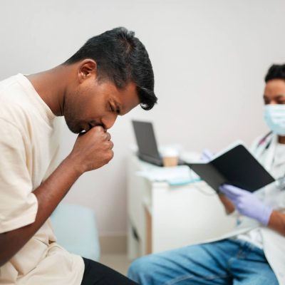 Man coughing during a medical consultation while a healthcare professional reviews patient notes